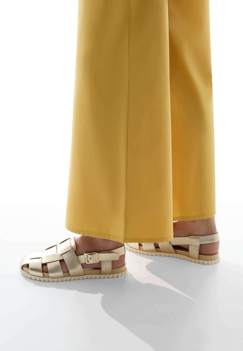 Sandales strappy beige clair avec détail à boucle, semelle texturée et tige tissée, associées à un pantalon jaune à jambes larges.
