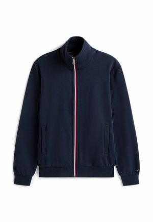 Veste de sport marine avec un col haut, fermeture éclair intégrale, détails en rouge et blanc le long de la fermeture éclair et poches latérales. Matériel doux.