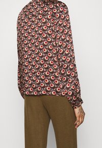 Blouse à manches longues avec un motif floral marron comprenant des fleurs rouges et blanches sur un fond sombre. Tissu léger avec une texture douce.