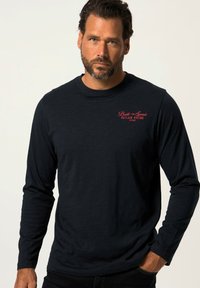 Camisa negra de manga larga en una tela suave que presenta un gráfico de texto rojo "Construido para la Velocidad Carreras Fuera de la Ley" en el área del pecho izquierdo.