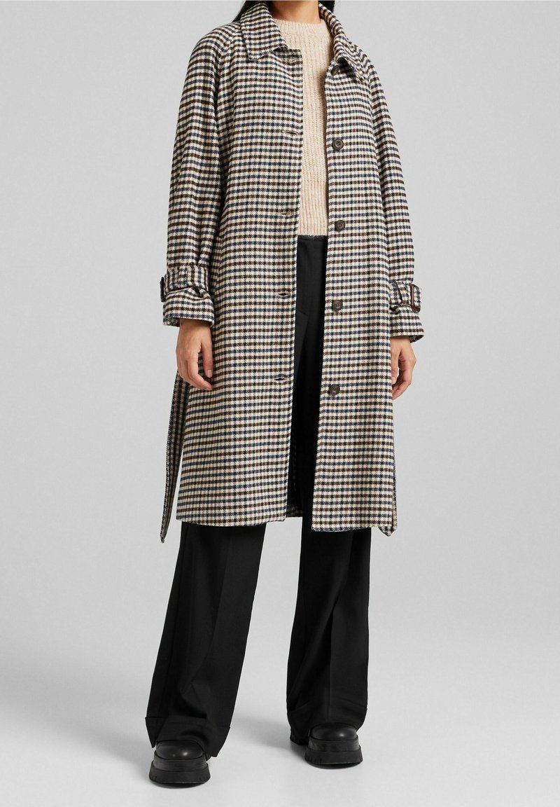 Femme portant un long manteau à carreaux avec des boutons, un pull beige, un pantalon noir à jambes larges et des chaussures à plateforme noires sur un fond gris uni.