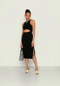 Vestido midi preto recortado com design sem mangas, fendas laterais e silhueta ajustada. Os acessórios incluem uma bolsa de padrão zebra e sandálias amarelas.