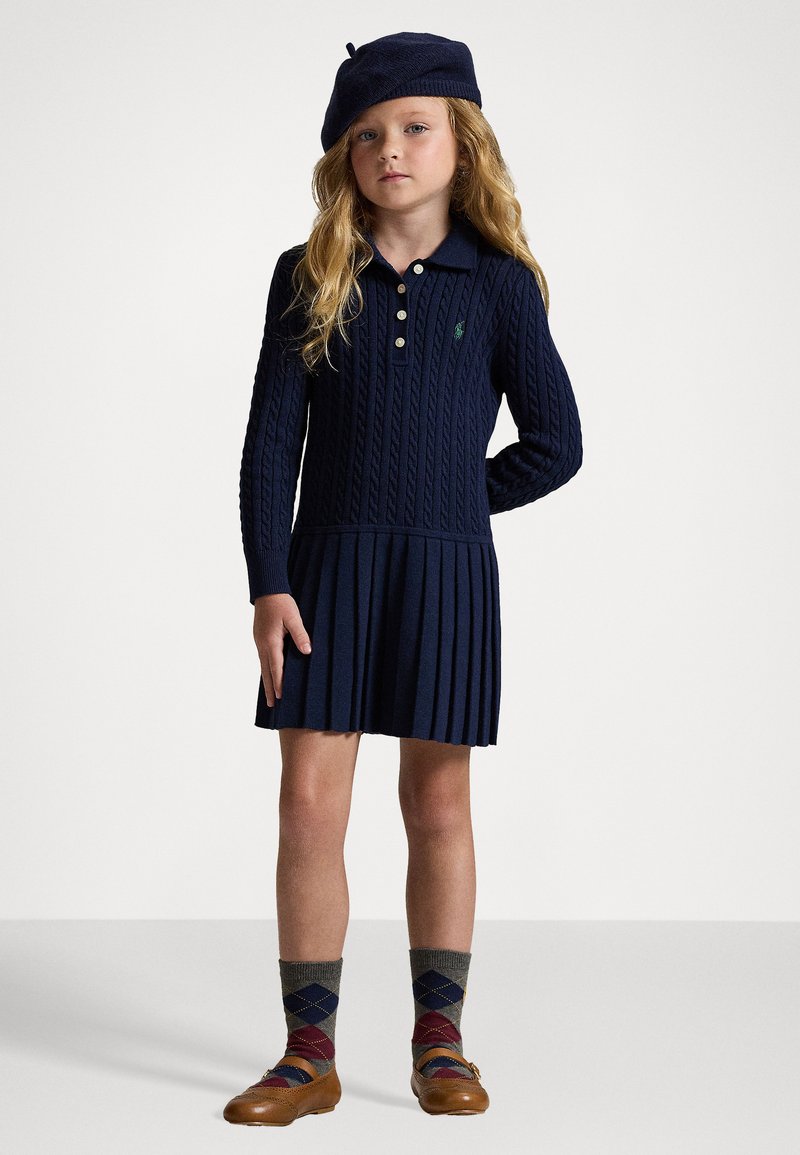 Mornarsko pleteno polo obleko z nabranim krilom, tremi gumbi in zelenim logotipom; kombinirana z beretko, argyle nogavicami in rjavimi čevlji.