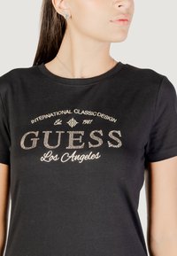 T-shirt in cotone nero con la scritta "GUESS" in strass argento, con "Los Angeles" in bianco in corsivo sotto, scollo tondo e maniche corte.