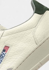Sneaker in pelle bianca texturizzata con dettaglio grigio sul colletto e linguetta verde sul tallone. Presenta una patch con logo e cuciture a contrasto.