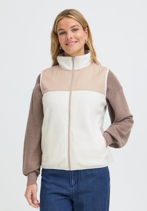 Femme souriante portant une veste sans manches beige et blanche zippée par-dessus un haut marron à manches longues et un jean bleu, sur fond blanc.