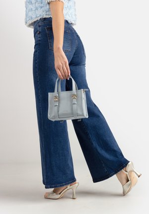 Donna che indossa jeans blu a gamba larga e sandali con tacco alto tiene una piccola borsa azzurro chiaro su uno sfondo semplice.