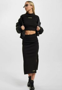 Svart ribbet crop topp med "ROCAWEAR" logo, parret med et matchende langt skjørt med en sideslitt. Modellen har på seg svarte joggesko og solbriller.