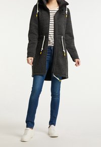 Kvinna iklädd en mörkgrå huvjacka med vita dragsnören, randig tröja, blå jeans och vita sneakers, som står mot en ljus bakgrund.