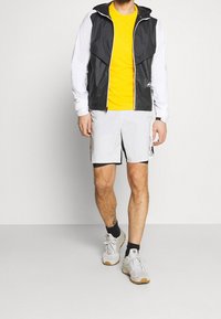 Vêtements de sport pour hommes : veste à capuche noire et blanche, t-shirt jaune, shorts gris clair avec des finitions noires, baskets blanches à semelles marron.