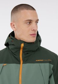 Grüne und graue wasserdichte Jacke mit hohem Kragen, orangefarbener Reißverschluss und Kapuze. Mit Logo und minimalistischem Design.