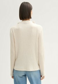 Femme avec les cheveux bruns courts portant un haut à manches longues texturé crème et un jean bleu clair, vue de dos sur un fond uni.