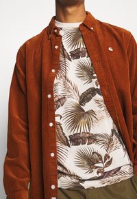 Mann in einem unknöpften, rostfarbenen Cordhemd über einem beigen T-Shirt mit grünen und braunen tropischen Blättern.
