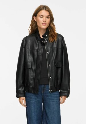 Femme portant une veste en cuir noire par-dessus un top noir avec un jean bleu, debout devant un fond blanc uni.