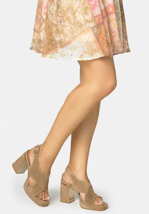 Mujer con sandalias de tacón ancho y punta abierta de ante beige y falda con estampado rosa y beige, de pie con una pierna cruzada sobre la otra.