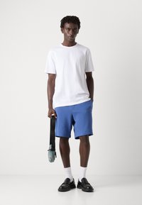 Tricou alb cu mâneci scurte, pantaloni șort albaștri, mocasini negri și șosete albe. Ține o geantă bleu deschis cu o curea neagră. Texturi de țesătură netedă.