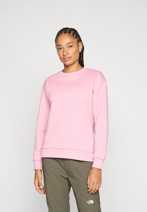 Donna con capelli intrecciati che indossa una felpa rosa chiaro e pantaloni verde oliva della North Face, in piedi contro uno sfondo chiaro e uniforme.