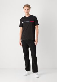 Schwarzes Baumwoll-T-Shirt mit einem weißen und roten horizontalen Streifen und dem Aufdruck "Tommy Hilfiger" in der Mitte, kombiniert mit schwarzen Jeans und weißen Sneakern.