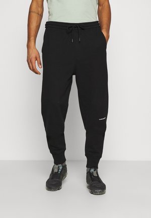 Mężczyzna ubrany w czarne spodnie dresowe z troczkiem w pasie, szare skarpetki i czarne buty sportowe, stojący na jednolitym tle.