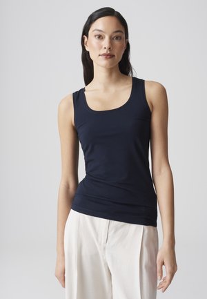 Femme aux longs cheveux foncés portant un débardeur bleu marine ajusté et un pantalon blanc ample sur un fond clair uni.