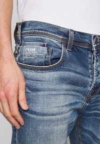 Vyblité modré džíny s rovnýcm střihem, s nášivkou na zadní kapse a kontrastním švem. Ruka v těsné blízkosti látky.