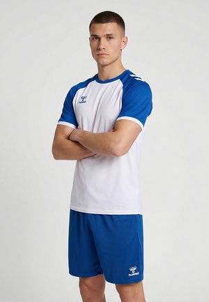 Młody mężczyzna z krótkimi włosami stoi z założonymi ramionami, ubrany w białą i niebieską odzież sportową z logo hummel na koszulce i spodenkach.