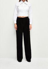 Camicia bianca crop con un ampio colletto e bottoni, abbinata a pantaloni neri a gamba larga. Il tessuto sembra liscio e ben strutturato.