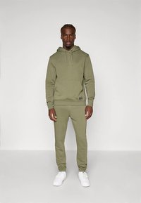 Σετ με φανελένιο φούτερ σε ελιά πράσινο και τζόγερς που ταιριάζουν, φτιαγμένο από απαλό ύφασμα με εμπρόσθια τσέπη, ριμπ τελειώματα και λευκά σνίκερς.