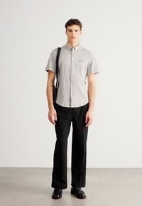 Szara koszula na krótki rękaw z guzikami, czarne spodnie i czarne buty. Model trzyma czarną torbę. Prosty wzór i klasyczny krój. Gładka tekstura.