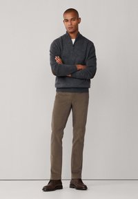 Pull gris à col zippé, chinos marron et chaussures marron foncé. Le modèle se tient les bras croisés, contre un fond neutre.