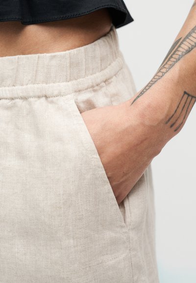 Μπεζ λινό σορτς με ελαστική μέση και πλαϊνή τσέπη, με υφή στο ύφασμα και χαλαρή γραμμή. Κοντινή εικόνα της περιοχής της τσέπης.
