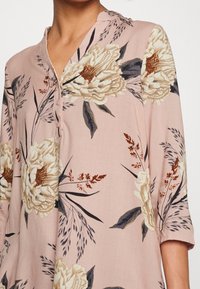 Rosa blus med v-ringning och trekvartslånga ärmar, dekorerad med stora beigea blommönster och mörkgröna lövverk.