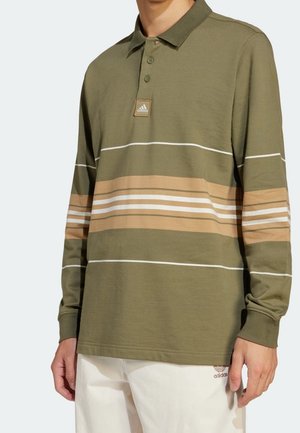Mężczyzna ubrany w oliwkowozieloną koszulkę polo z długim rękawem, z beżowymi i białymi poziomymi paskami oraz logo Adidas, w połączeniu z białymi spodniami Adidas.