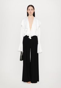 Witte ruches blouse met diepe V-hals, gecombineerd met zwarte wijde broek. Model houdt een kleine zwart gestructureerde handtas vast.