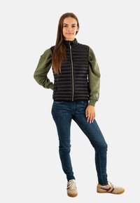 Chaleco acolchado negro sobre una camiseta de manga larga verde, con cierre de cremallera. Combinado con jeans azules y zapatillas beige con acentos en marrón.