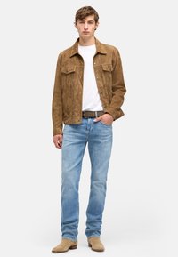 Jeune homme portant une veste en suède marron, un t-shirt blanc, un jean bleu clair, une ceinture marron et des bottes en suède beige, debout avec une main dans la poche.