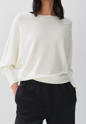 Femme portant un haut blanc ample à manches longues et un pantalon noir, les mains dans les poches, sur un fond gris uni.