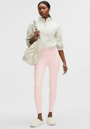 Frau trägt ein cremefarbenes Sweatshirt mit Viertel-Reißverschluss, hellrosa Leggings, Slipper und trägt eine große cremefarbene Schultertasche mit Rüschen.