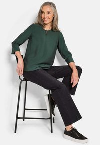 Eine Frau mit grauen Haaren sitzt auf einem schwarzen Hocker und trägt ein dunkelgrünes Langarmoberteil, schwarze Hosen und schwarze Sneakers mit weißen Sohlen.