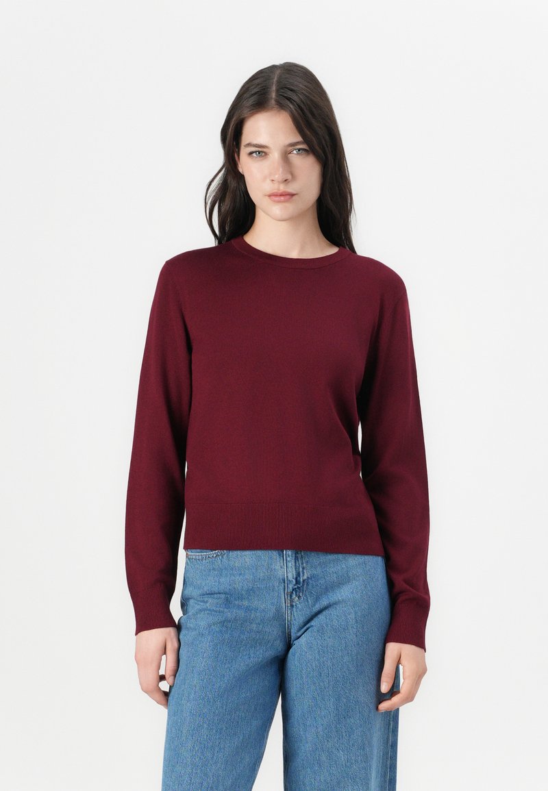 Burgundska pletena pulover s okroglo ovratnico in dolgimi rokavi, ki ima prilegajoče se pas in rebraste manšete, v kombinaciji s svetlo modrimi jeans hlačami.