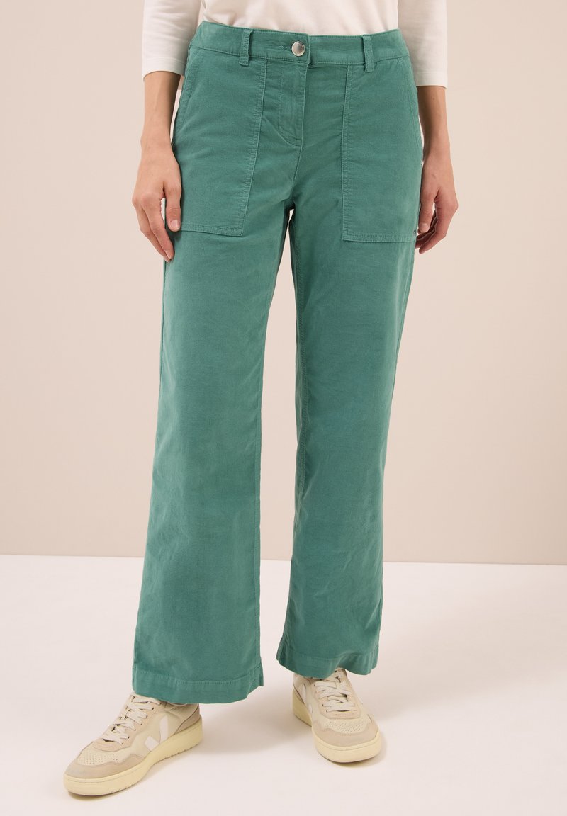 Zelené kalhoty s širokými nohavicemi vyrobené z měkké tkaniny, s předními kapsami a zapínáním na knoflík v pase, v kombinaci se světlými teniskami.