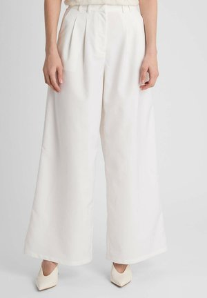 Vrouw draagt witte hoge taille wijde pijpen met plooien, gecombineerd met witte puntige hakken, staande tegen een effen achtergrond.