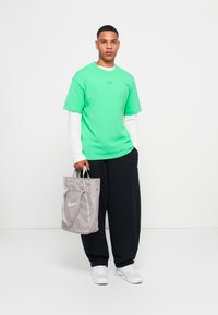Tricou verde din bumbac cu un logo, pus peste o cămașă albă cu mâneci lungi. Pantaloni de trening negri și o geantă model gri cu detalii de logo. Adidași albi.