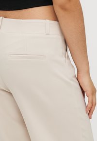 Pantaloni beige chiaro con una texture liscia, vestibilità sartoriale e una tasca posteriore singola. Le cuciture laterali creano una silhouette slanciata.