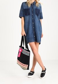 Vestido de camisa de denim con mangas cortas, dos bolsillos en el pecho; bolsa de tote negra con bordado colorido y borlas rosas; zapatillas negras con acentos de perlas.