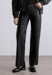 Μαύρο δερμάτινο παντελόνι με ψηλή μέση, με κάθετες ραφές και ίσια γραμμή. Συνδυάζεται με μαύρα γυαλιστερά loafer.