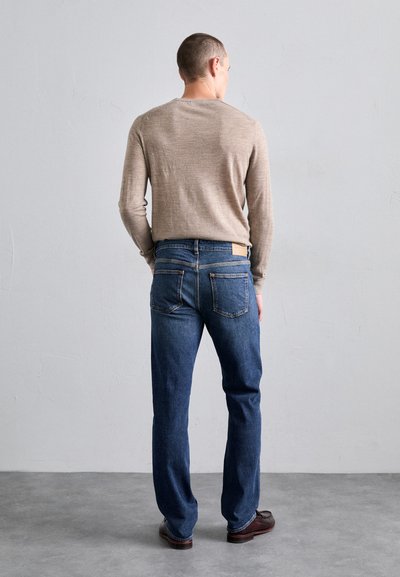 Une chemise beige à manches longues avec un col rond, associée à un jean bleu foncé. Le jean présente une patch en cuir à l'arrière et des poches visibles.