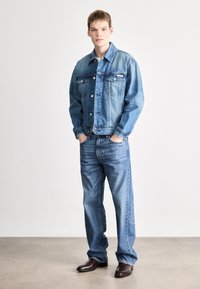 Τζιν μπουφάν και τζιν παντελόνι σε ανοιχτό μπλε. Το μπουφάν έχει μεγάλες μπροστινές τσέπες, κουμπιά και άνετη γραμμή. Τα καφέ δερμάτινα παπούτσια ολοκληρώνουν την εμφάνιση.