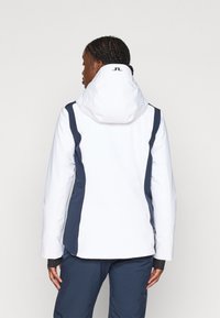 Balta striukė su tamsiai mėlynomis šoninėmis juostomis, aukštu apykakle ir kapa. Yra minimalizmo dizainas su logotipu nugaros dalyje. Lygi tekstūra.