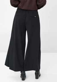 Pantalons larges noirs en tissu texturé, avec une taille froncée et deux poches arrière, associés à des bottes noires à talons.